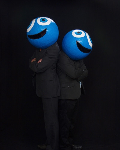 The Bran Flakes once again donning smiling masks, but this time they have their backs against each other and their arms crossed. They are in a black background.